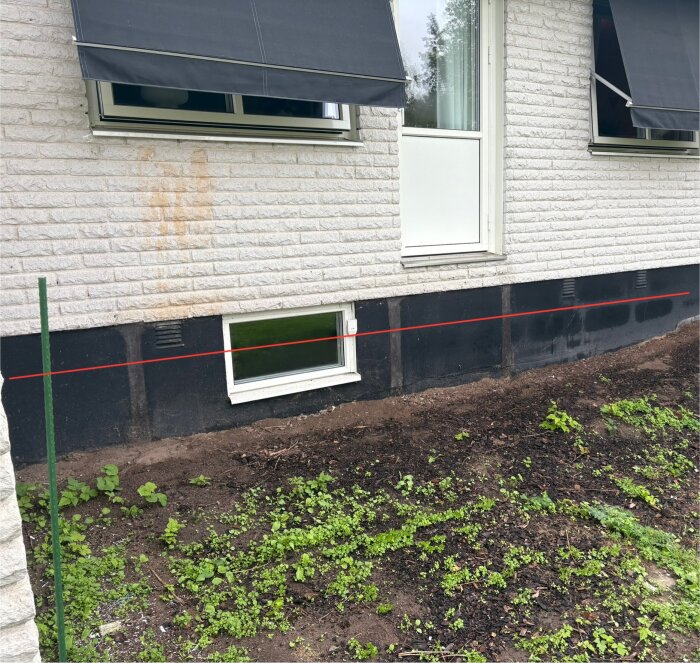 Fasad på ett hus med fönster och en dörr. Ett rött streck markerar höjden för planerad altan. Jord och växtlighet syns nedanför fönstret.