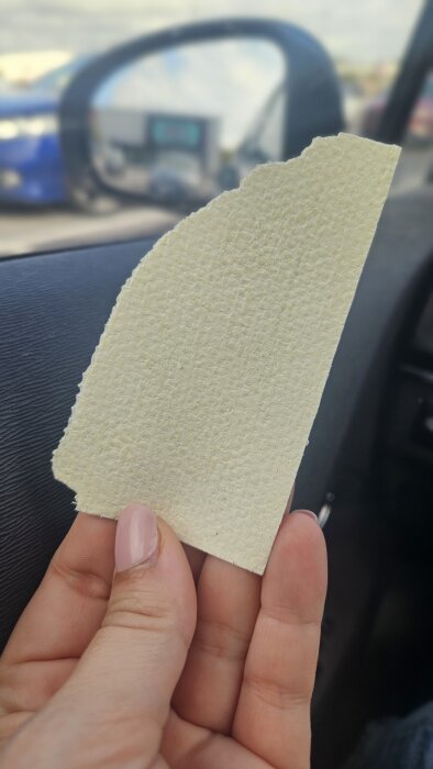Hand håller ett ljusbeige provbit av väv med tydlig struktur, mot en bilinteriör.