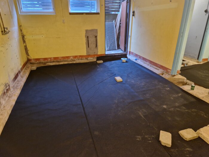 Rum med svart markduk på golvet, beredd för gjutning, med stenar och öppet fönster, vid förberedelse för installation av golvvärme.