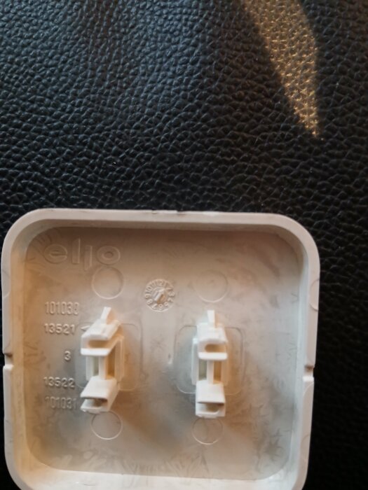 Lock av plast med två fästen, texten "elio" och olika nummer tryckta, ligger på en mörk yta.