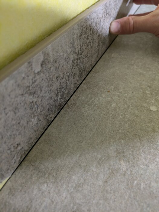 Hand justerar en sockellist mot klinkerplattor i badrum, med liten springa emellan.