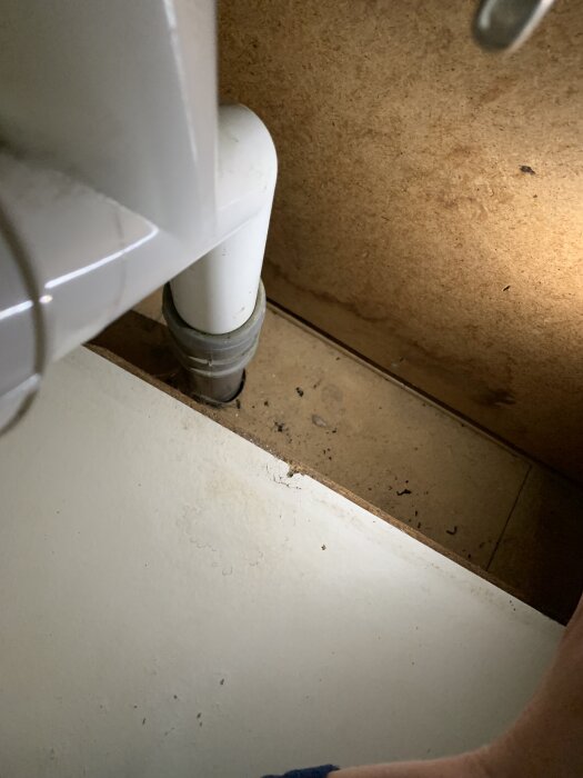 Vaskens undersida med vattenrör, potentiellt läckage synligt på grund av våtfärgade fläckar och smuts på träskivan.