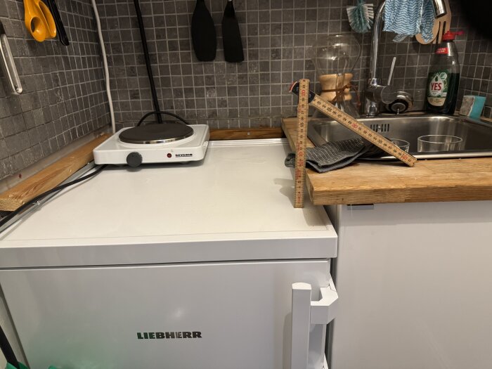 Kylskåp med måttstock och induktionsplatta ovanpå, bredvid träskiva och diskho i kök med mörkt kakel.