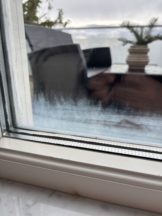 Otydlig beläggning mellan två fönsterrutor med utsikt över en uteplats och krukväxt, ingen synlig kondens eller fukt.