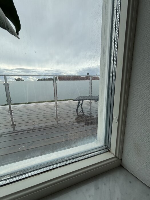 Utsikt genom fönster med något vitt mellan glasen, synlig veranda och molnig himmel utanför.