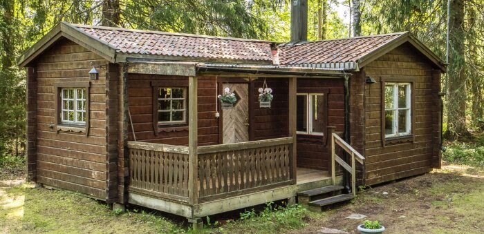 Trästuga i skogen med en liten veranda och blomsterrabatter, omgiven av träd.