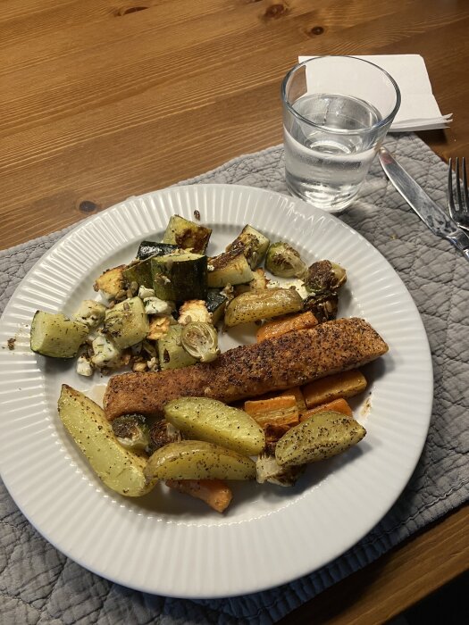 Tallrik med stekt lax, zucchini, solrosfrön, citron, dill, ugnsrostad potatis, morot och brysselkål. Glas med vatten och bestick bredvid.