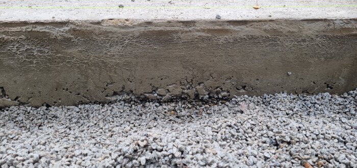 Betongkant med grov, ojämn yta och synlig ballast, intill grusunderlag.
