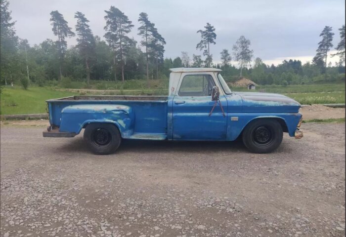 Blå äldre pickup med rostfläckar på karossen står parkerad på en grusväg, omgiven av träd och grönska.