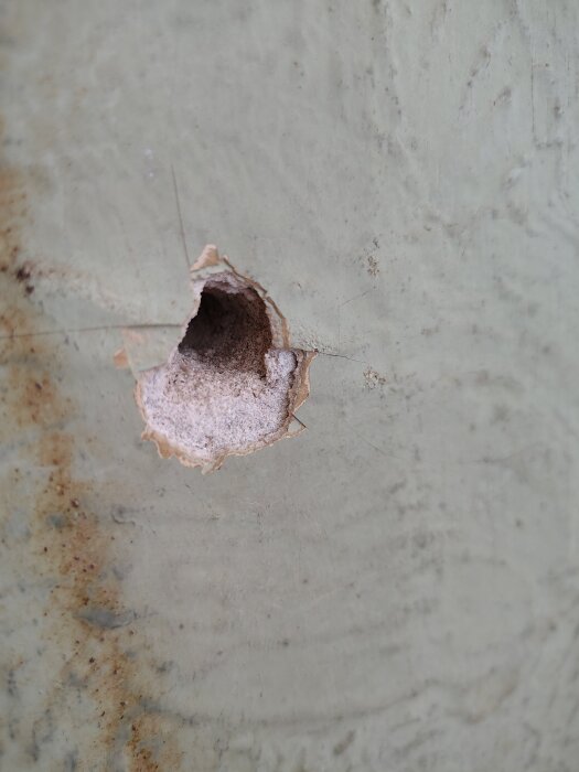 Gammal vägg med hål exponerar gipslager, ursprunglig färg syns. Tecken på rost och slitage synliga runt hålet.