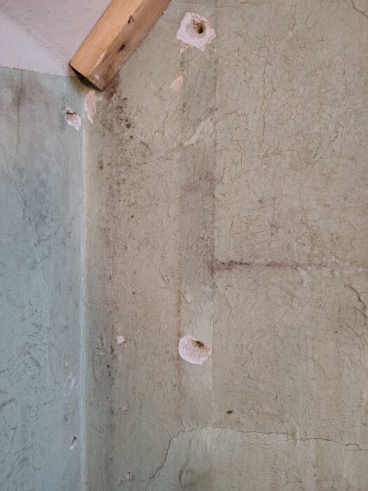 Sliten vägg i trapphus med sprickor och avflagningar, delvis exponerad rå puts och synlig trälist i hörnet.