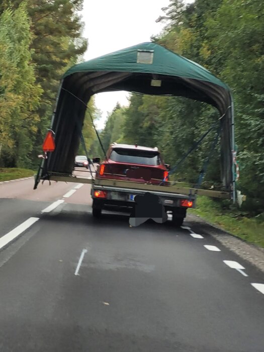 Bil med stor last av presenning och LGF-skylt på landsväg omgiven av skog.