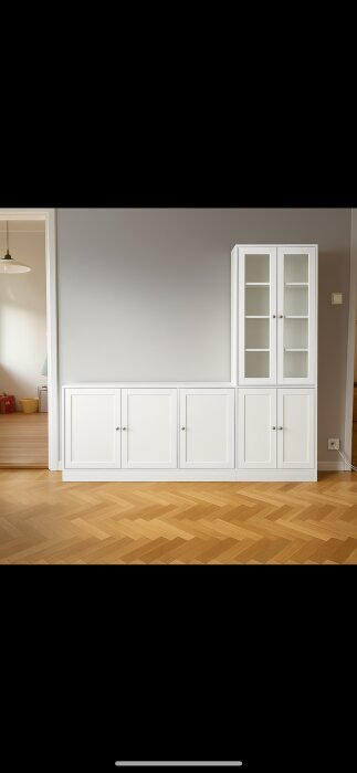 Vit väggfast skåpsenhet med fyra paneldörrar och glasdörrar på ena sidan, stående mot en grå vägg med trägolv.