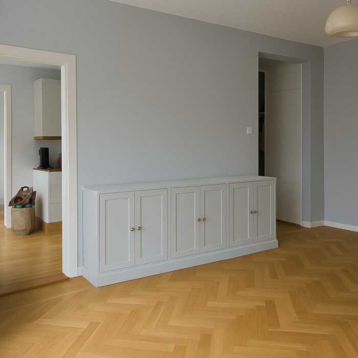 Grå vägg med en platsbyggd vit skänk längs golvet i ett rum med parkettgolv och dämpad belysning.