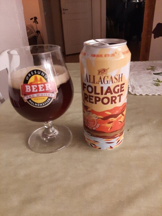 En glas med mörk ale och en burk med Allagash Foliage Report öl placerade på ett bord.