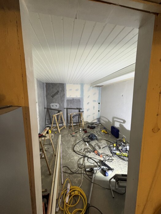 Rum under konstruktion med pärlspontstak och verktyg spridda på golvet, inklusive slagborr och kablar. Arbetsbänkar i bakgrunden.
