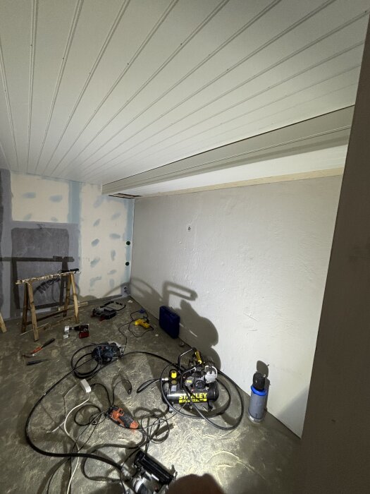 Renoveringsprojekt med pärlsponttak och verktyg på golvet, inklusive kompressor och såg. Vägg med gipsfinish i bakgrunden.