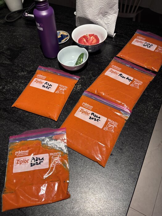Ziploc-påsar med hemgjord pizzasås markerade "Pizza 2025", tomat i skål och lila vattenflaska på köksbänk.