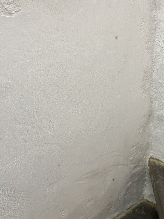 Vägg med vit putsytan där små mörka fläckar har uppstått efter målning, synliga nära hörnet och spridda över ytan.