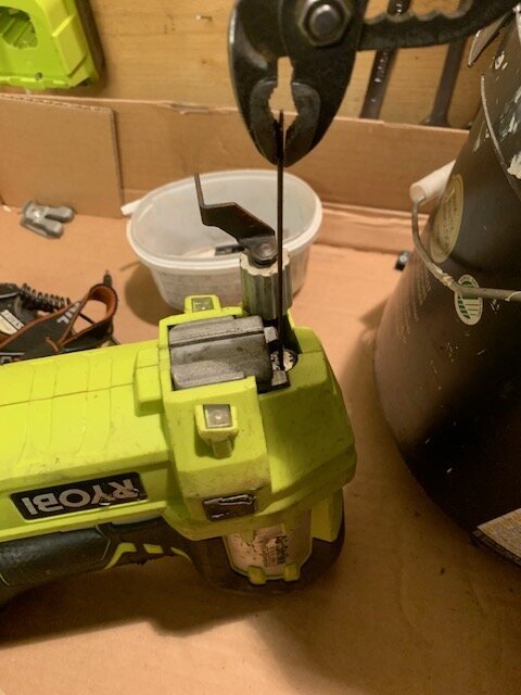 En grön Ryobi spikpistol med ett verktyg insatt i toppen, omgiven av en plastburk och en hink, på en arbetsyta.