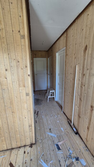 Renoveringskaos i hall, masonitväggar och råspont syns. Golvet har skräp och stegpall står vid stängd dörr. Reglar ska riktas för jämnhet.