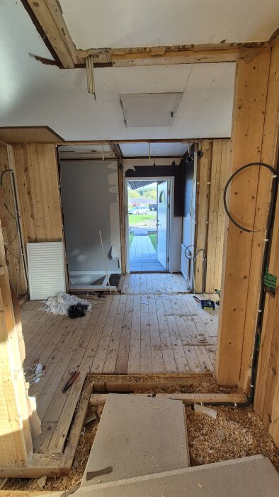 Renovering av interiör med synligt trä och rör, öppen dörr mot utsidan, halvfärdigt golv. Verktyg och byggmaterial ligger på plats.
