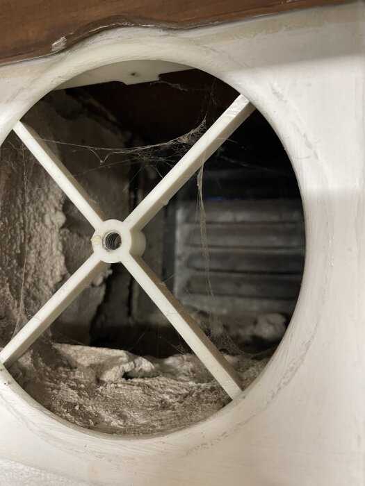 Ventilationskanal med plastgaller täckt av damm och spindelväv, sett inifrån ett trångt utrymme.