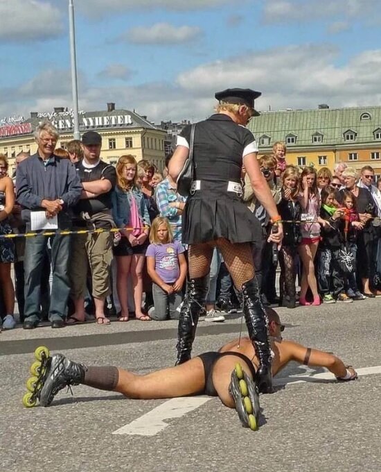 Person i svart uniform och högklackade stövlar står över en liggande person med rullskridskor på gatan, omgiven av publik.