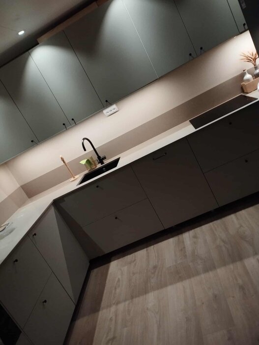 Modernt kök med gråa skåp, ljus bänkskiva, svart kran och spis, samt trästavar på golvet.