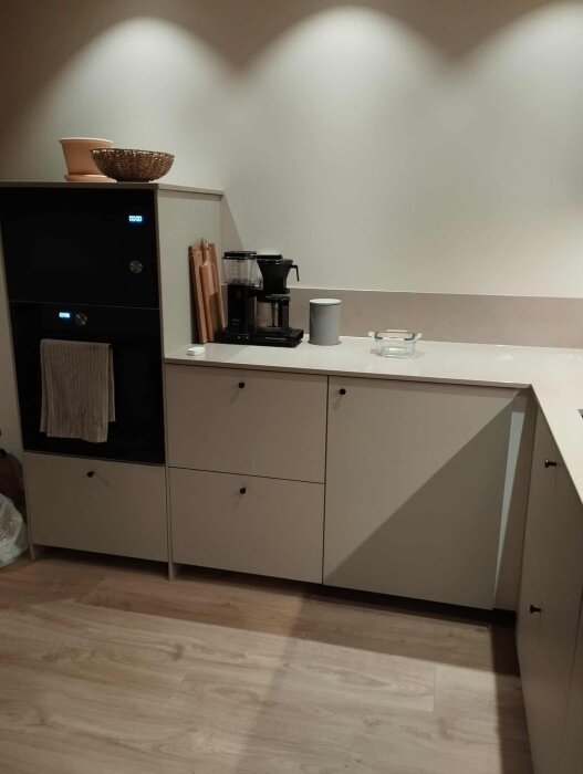 Modernt kök med ljusa skåp, inbyggd dubbelugn, kaffebryggare och skärbrädor. Trägolv och minimalistisk inredning.