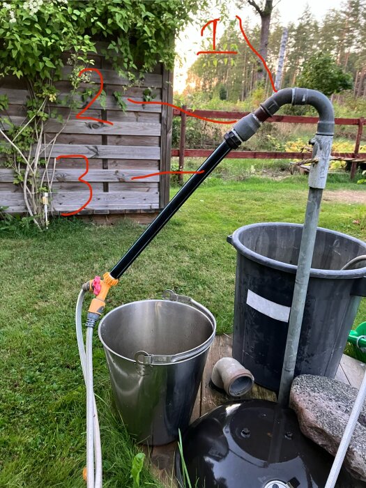 Rör med markeringar i trädgård, tre kopplingar i metall, en hink och en plastslang, mot en träribbad vägg och gräsmatta.