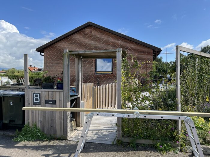 Entréportal under renovering framför tegelhus, med brädor, stege och grönska i bakgrunden samt blå himmel.