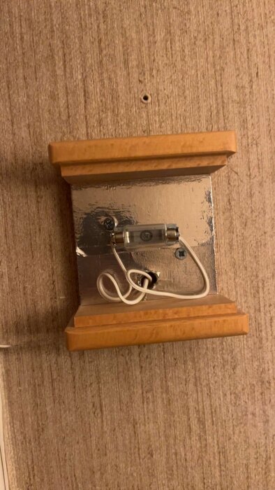 Gammal ringklocka kopplad till en lampa, monterad på en trälist med exponerade kablar och en metallplatta på väggen.