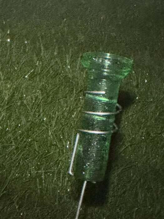 Grön glascylinder med en pinne, på mörk yta, fylld av regnvatten.