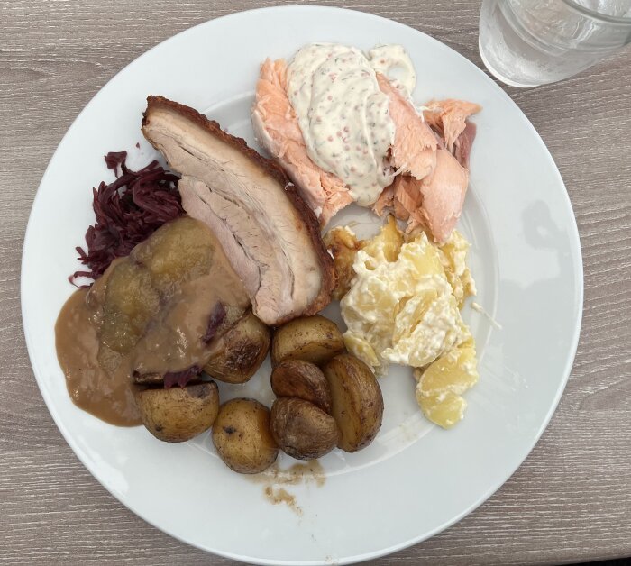 Tallrik med fläskstek, lax, potatisgratäng, små potatisar, rödkål och sås på ett bord.