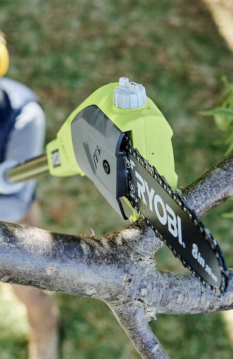 En grön Ryobi grensåg använd utomhus. Sågen vilar mot en trädgren.