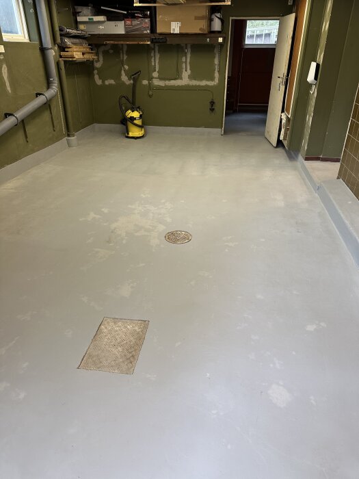 Matt och flammigt garagegolv målat med epoxifärg, fläckvisa blanka partier. Väggar och hyllor i bakgrunden, dammsugare på golvet.