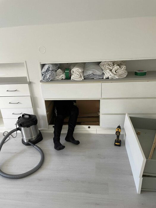 Person inspekterar bakom platsbyggd byrå på en övervåning, med dammsugare och verktyg nära, i rum med ljusa väggar och golv.
