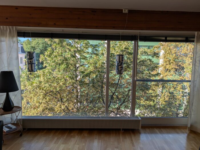 Stort fönster med träbjälke ovanför och radiator längs golvet, utsikt mot lummiga träd och byggnader utanför, golvlampa till vänster.