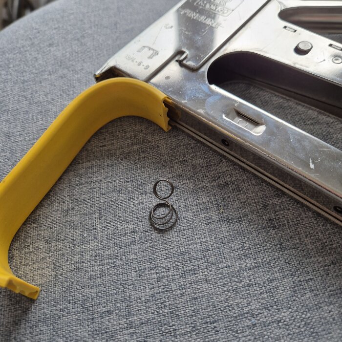 Trasig häftpistol med borttagen gul plast och deformerad fjäder på grå yta.