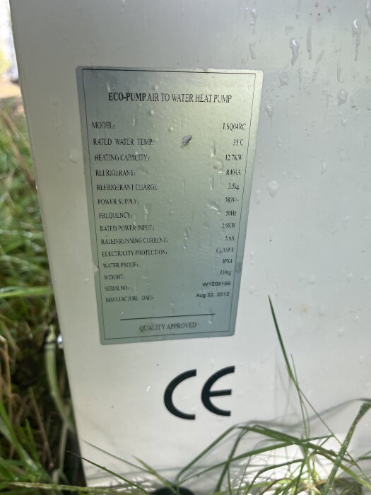 Informationstavla på en luft-vatten-värmepump, med tekniska specifikationer och CE-märkning, omgiven av gräs.