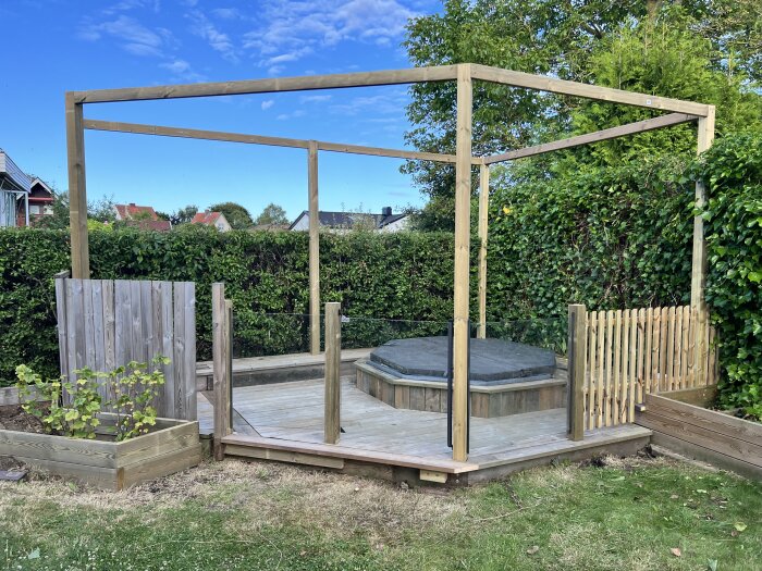 Träpergola med glas runt en upphöjd badtunna, omgiven av häckar och trästaket i en grönskande trädgård.