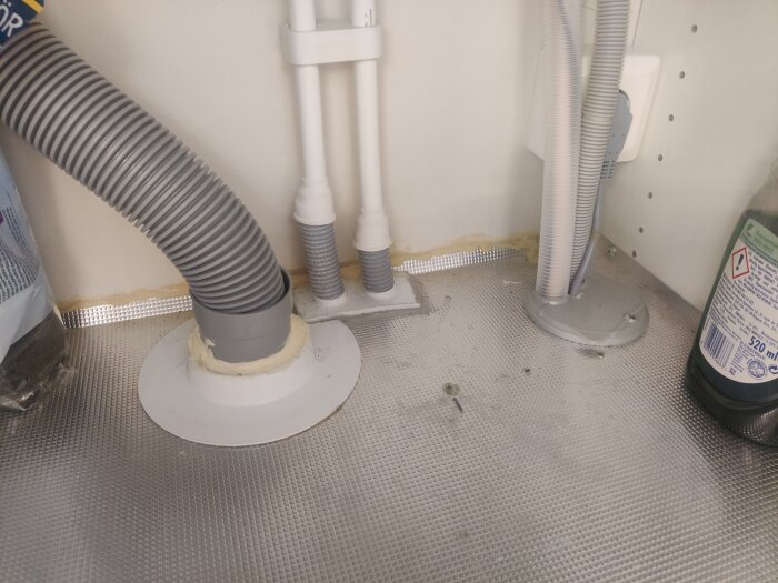 Grå slangar och rör under diskbänk, vit vägg och metallaktig yta, diskmedelsflaska syns i hörn.