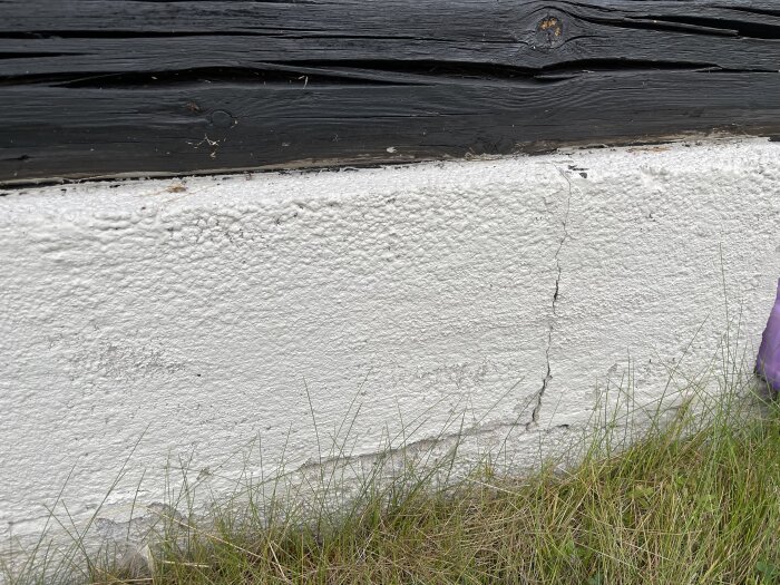 Vitmålad grund med spricka och svart träpanel ovanpå, omgiven av gräs.