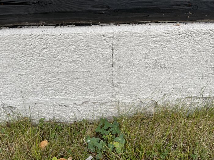 Målade murblock med sprickor och slitage vid basen, delvis täckta av gräs, på en äldre byggnad.