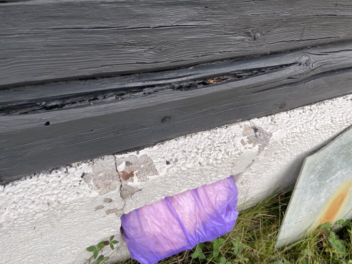 Sprickor i husgrund av betong med synliga lagningar, lila plastpåse i öppning. Svartmålad knuttimrad stockvägg ovanför. Matta grässtrån bredvid.
