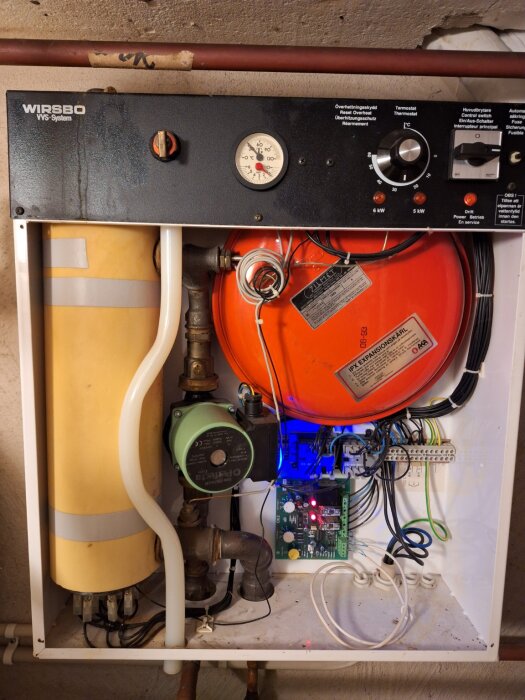 Fördelare för vattenburen golvvärme med tryckmätare, termostat och rörkopplingar inuti ett köksskåp, en del av ett äldre system.