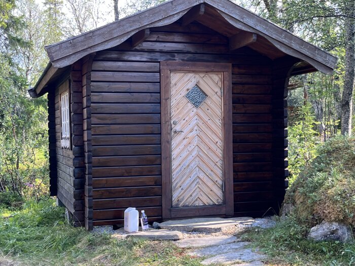 En timrad stuga med mörkbrun fasad, där dörren är ljusare och har ett chevronmönster. Stugan står i en skogsmiljö.