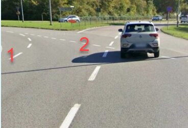 En bil kör i en cirkulationsplats. Vägen har två filer, den vänstra filen delar sig i två. Vägskyltar och träd syns i bakgrunden.