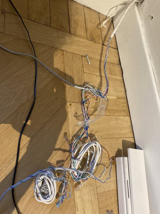 En trasslig bunt av kablar, inklusive vita och flerfärgade kablar, ligger på ett trägolv intill en vägg.